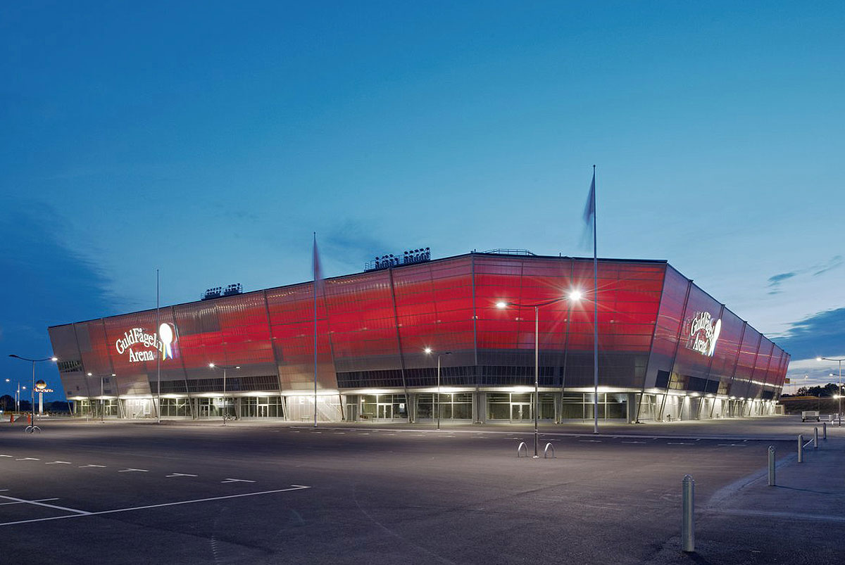 Guldfågeln Arena i Kalmar, i kvällsljus med en rödlysande fasad, omgiven av en tom parkeringsplats och blå kvällshimmel. Formentas flaggstänger står upplysta framför arenan.