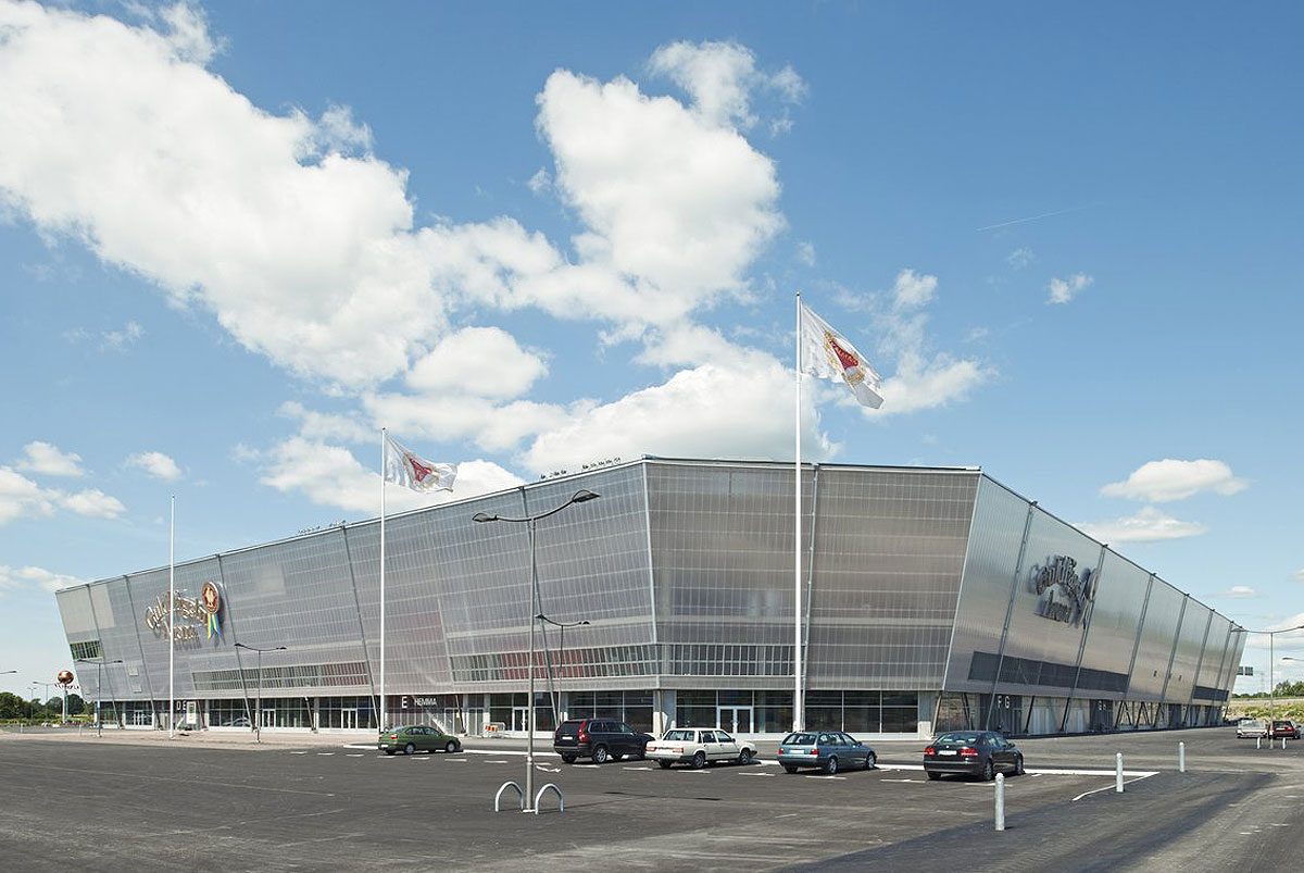 En vy av Guldfågeln Arena i dagsljus med blå himmel och vita moln, där två svenska flaggor vajar på höga flaggstänger från Formenta framför arenans moderna arkitektur. Bilar är parkerade framför byggnaden.