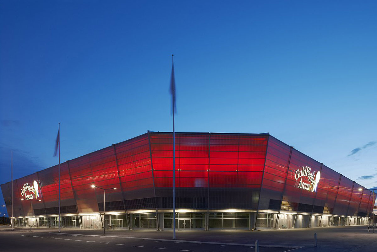 Guldfågeln Arena lyser upp i mörkret med sin karakteristiska röda belysning, omgiven av flaggstänger från Formenta som skapar en ståtlig siluett mot den blå natthimlen.